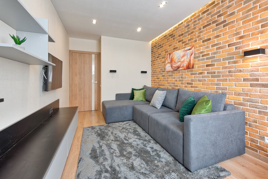 Interior view of a modern living room with a grey fabric sectional sofa adorned with green, blue, and grey cushions, positioned against a exposed red brick wall illuminated by black wall-mounted sconc