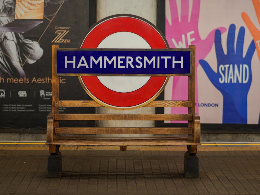 A wooden bench positioned on a tiled platform at the Hammersmith underground station, with a large round sign featuring a red circle and blue bar displaying 'HAMMERSMITH' in white letters mounted behi