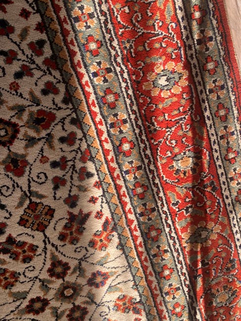 Close-up view of a traditional patterned rug with intricate floral and geometric designs in shades of orange, blue, and cream, covering a wooden floor in a well-lit dining area. A black metal chair wi