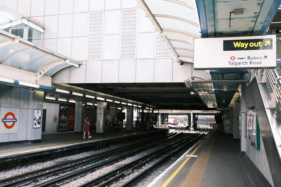 A wooden bench with black metal supports is positioned on a tiled floor in an underground station. Behind the bench, there is a large circular red and white sign with a blue rectangular sign overlayin